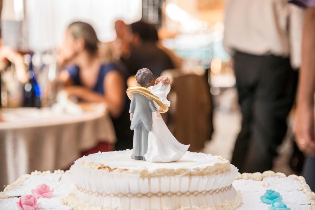 Nahaufnahme eines Hochzeitskuchens mit einer Figuren von Braut und Bräutigam als Topper, bei einer freien Trauung im Garten, mit unscharfen Gästen im Hintergrund