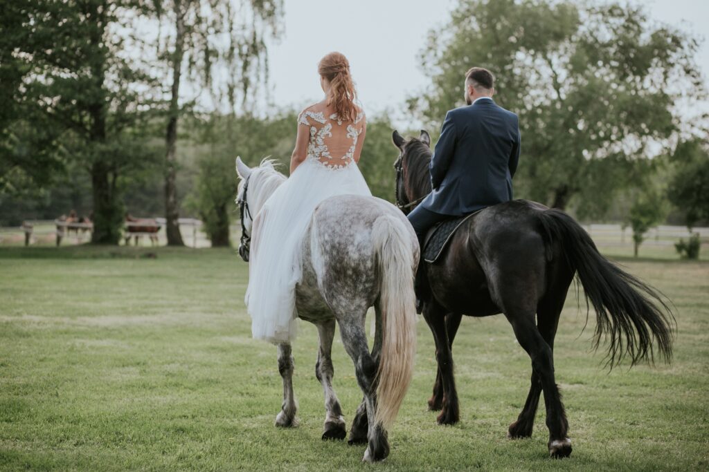 Brautpaar reitet auf Pferden während ihrer freien Trauung im Garten, die Braut in einem weißen Spitzenkleid und der Bräutigam in einem dunklen Anzug, umgeben von grüner Landschaft