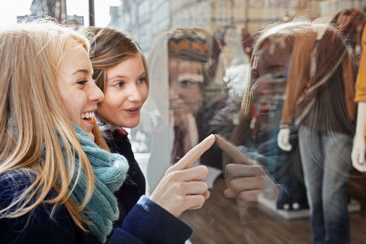 Zwei Frauen schauen in ein Mode-Schaufenster. Reflexion zeigt Jacken und Kleidung. Ist die John Doe Jacke eine sinnvolle Wahl?