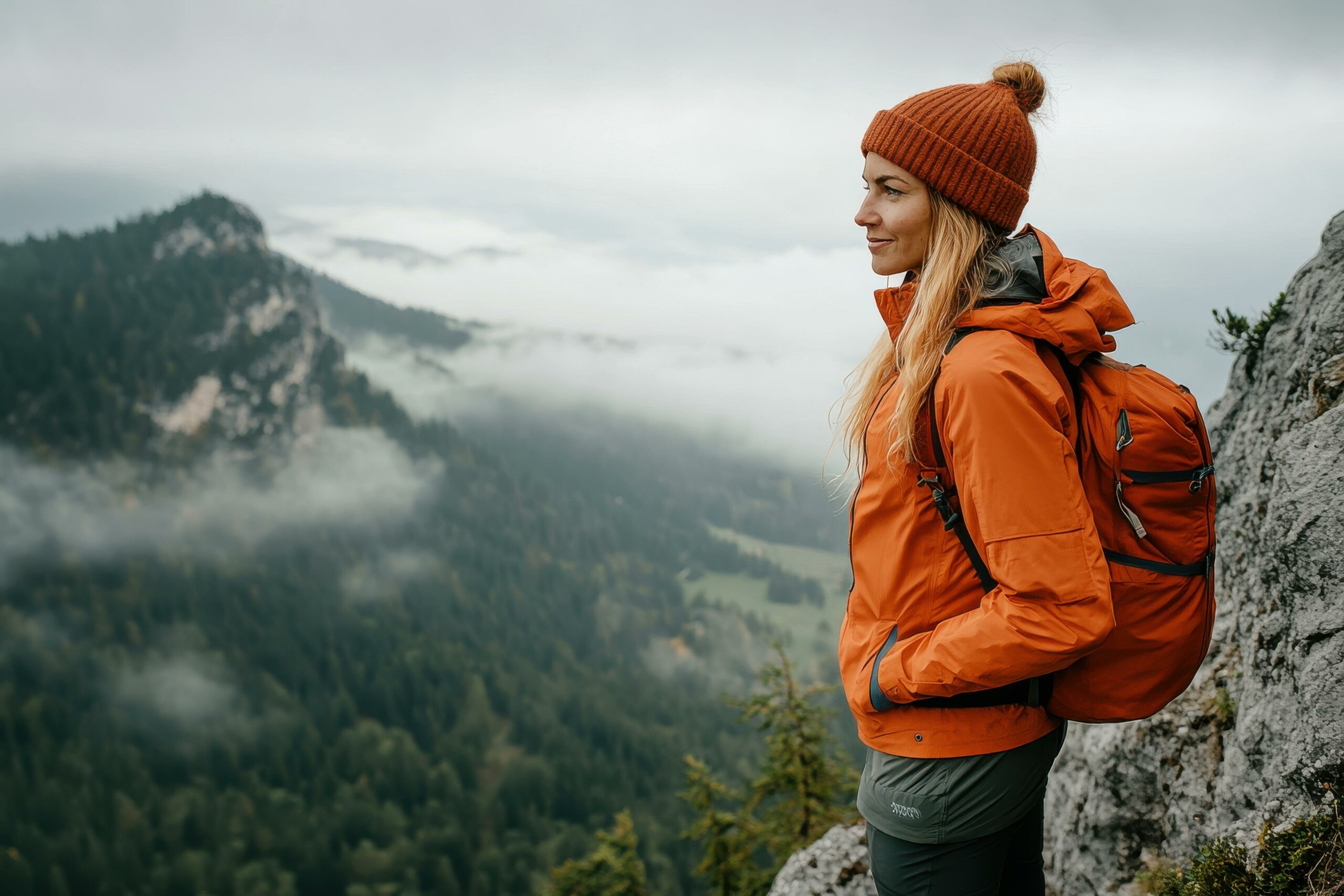 Frau mit John Doe Jacke und Rucksack steht in den Bergen. Multifunktionale Kleidung für Outdoor, Reisen und Alltag. Perfekt für wechselhaftes Wetter.